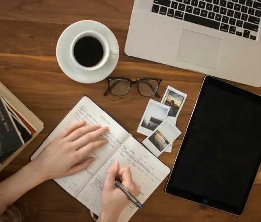 Hand writing in a notebook beside a laptop, tablet, coffee cup and glasses on a wooden desk, seen from above