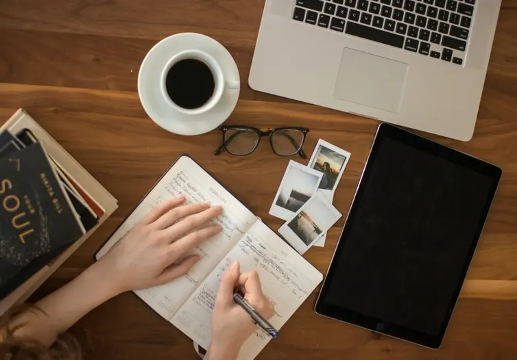 Hand writing in a notebook beside a laptop, tablet, coffee cup and glasses on a wooden desk, seen from above