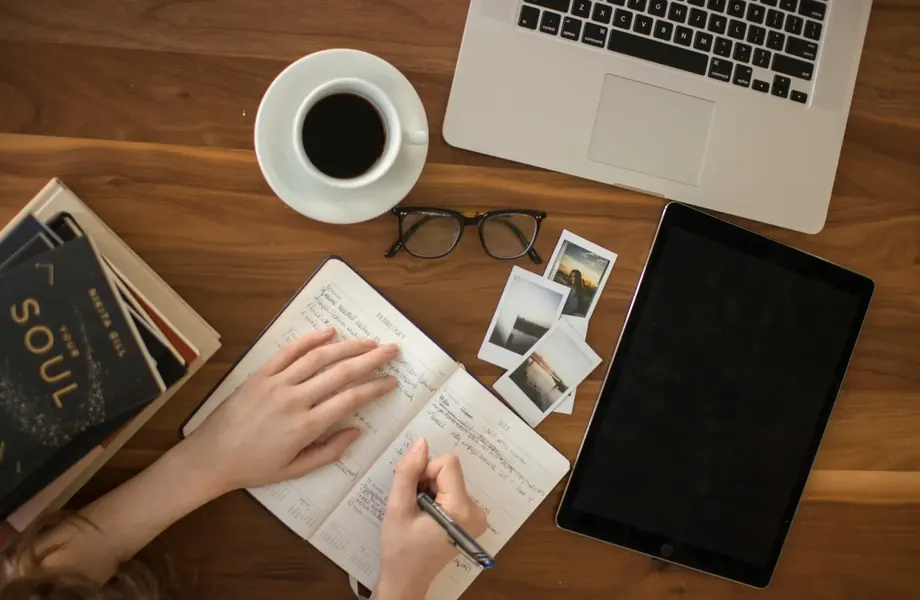 Hand writing in a notebook beside a laptop, tablet, coffee cup and glasses on a wooden desk, seen from above