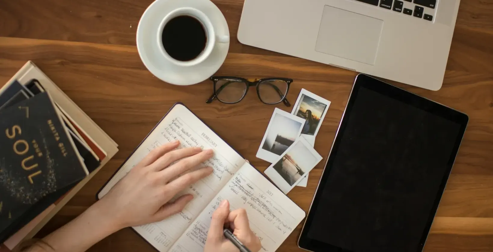 Hand writing in a notebook beside a laptop, tablet, coffee cup and glasses on a wooden desk, seen from above